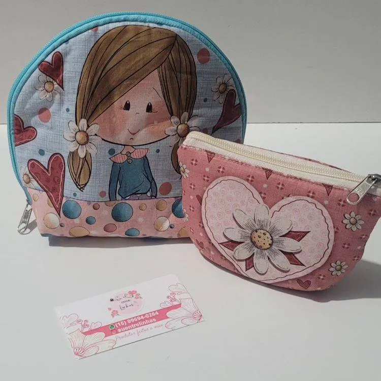 Imagem de uma necessáire em tons de azul com o desenho de uma menina e outra menor em tons de rosa. Próximo a elas, há um cartão da loja de Suzane.