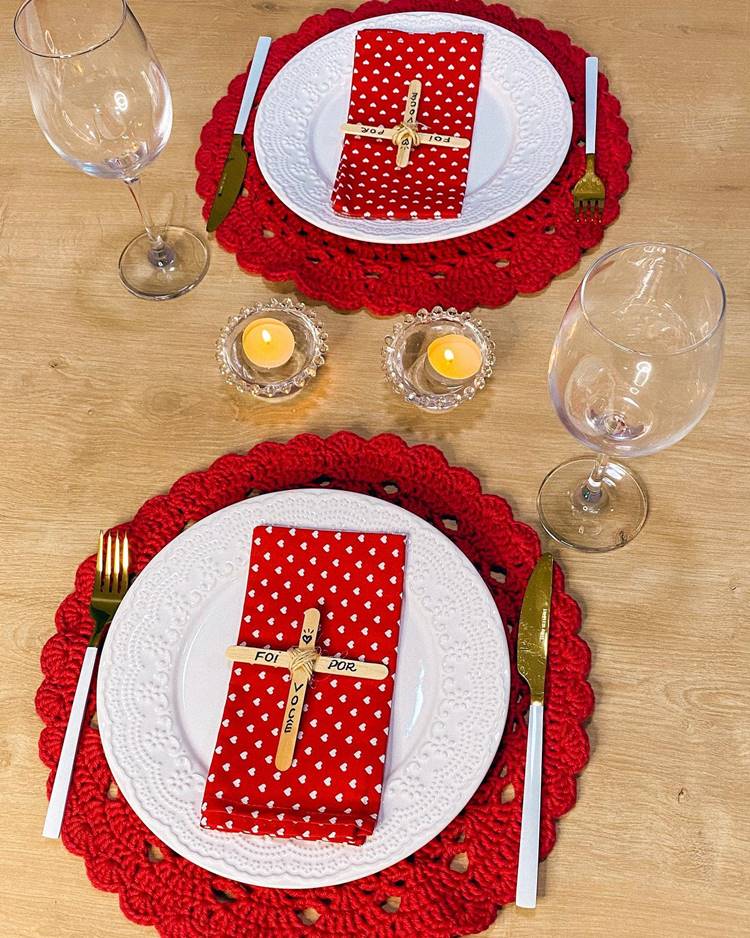 Uma mesa de jantar decorada para a Páscoa Cristã: sousplat vermelho de crochê, prato branco de cerâmica, guardanapo vermelho com estampa de corações, cruz feita de palito de picolé com a frase "Foi por você", talheres dourados, taças de vidro e vela.