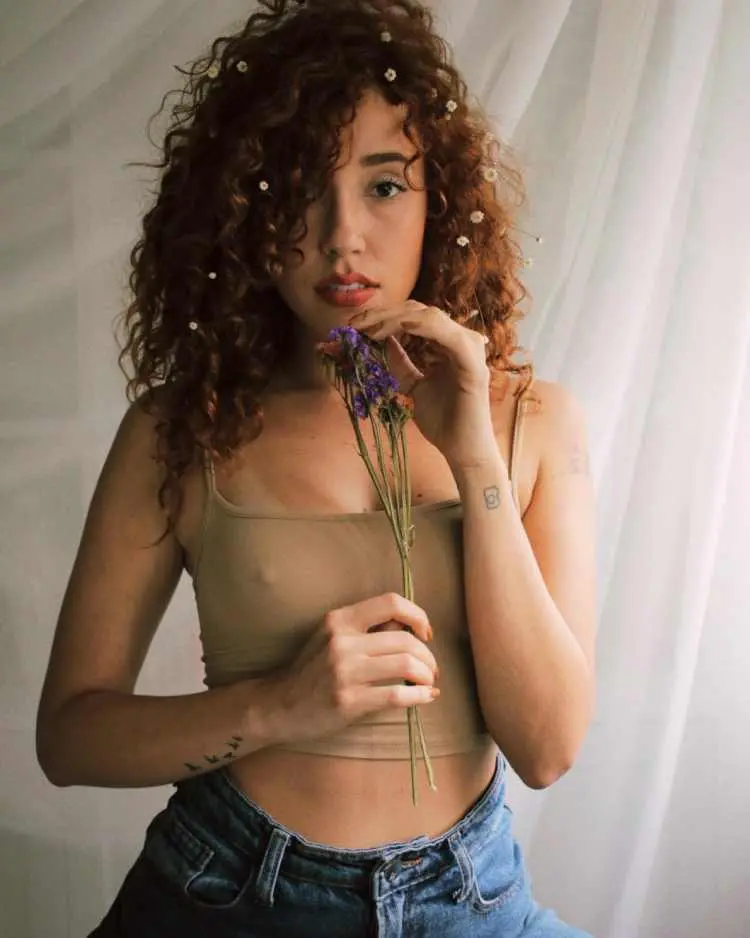Bruna Vieira com flores pequenas no cabelo, vestindo regata verde e calça jeans, está segurando flores roxas e está em fundo branco. É possível ver suas tatuagens no pulso: pássaros em um e uma câmera no outro