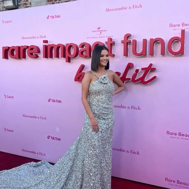 Selena Gomez posando em um tapete vermelho, ela usa um vestido de gala longo prateado e cabelo liso blunt reto