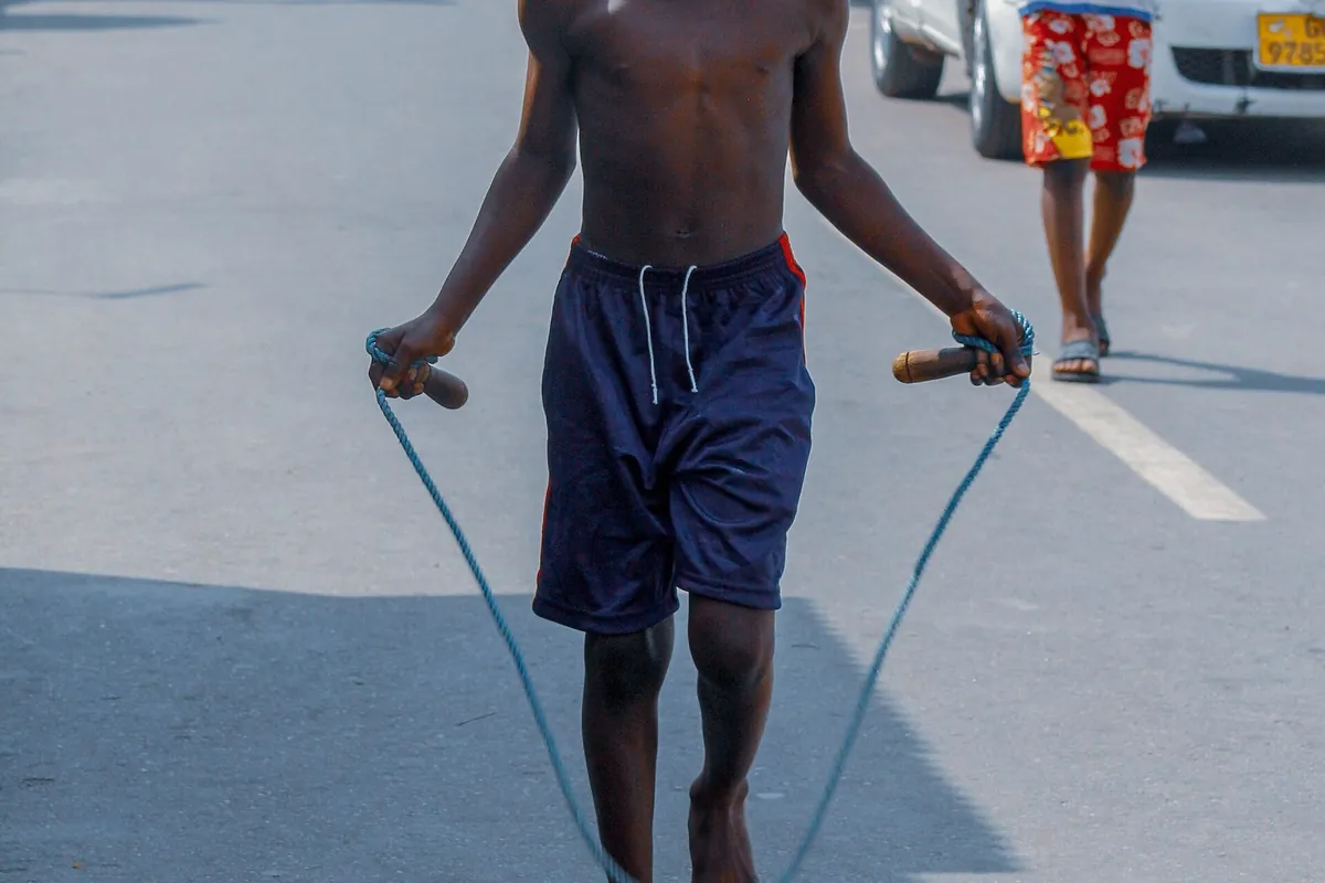 Jovem pulando corda em rua asfaltada, segurando os cabos com os cotovelos perto do corpo