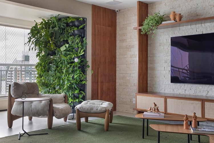  sala de estar moderna e bem iluminada. Há uma cadeira confortável e um pufe correspondente, ambos em um tecido cinza claro, no centro da imagem. Uma mesa de café moderna com tampo de vidro está à frente da cadeira. À esquerda, há uma exuberante parede viva com várias plantas verdes. À direita, uma TV plana está montada em uma parede de tijolos brancos. Um móvel baixo de madeira clara está sob a TV, adornado com alguns objetos decorativos e livros. O piso é coberto por um tapete verde que complementa as plantas na parede viva. A sala é espaçosa e tem janelas grandes que permitem a entrada de muita luz natural.