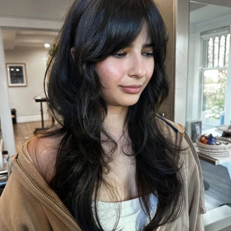 Uma mulher branca de cabelos longos, repicados e pretos, usando uma blusa branca e um casaco marrom