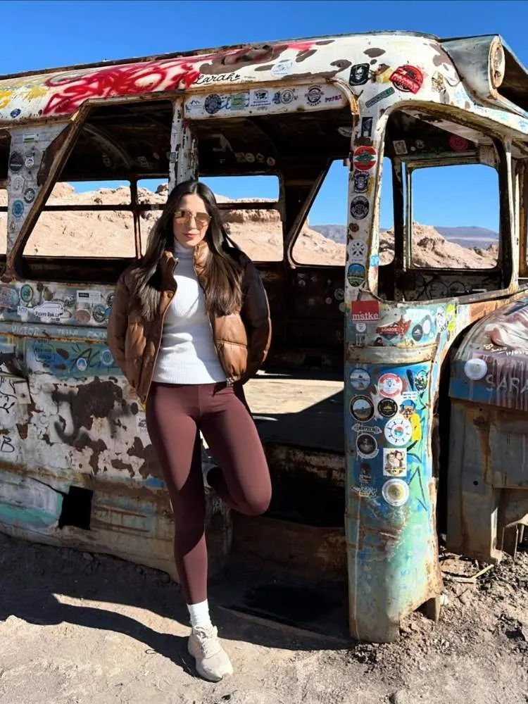 Mulher de pele clara apoiada em ônibus abandonado, usando cacharrel branca, puffer marrom, legging marrom, meia branca cano médio e tênis esportivo 