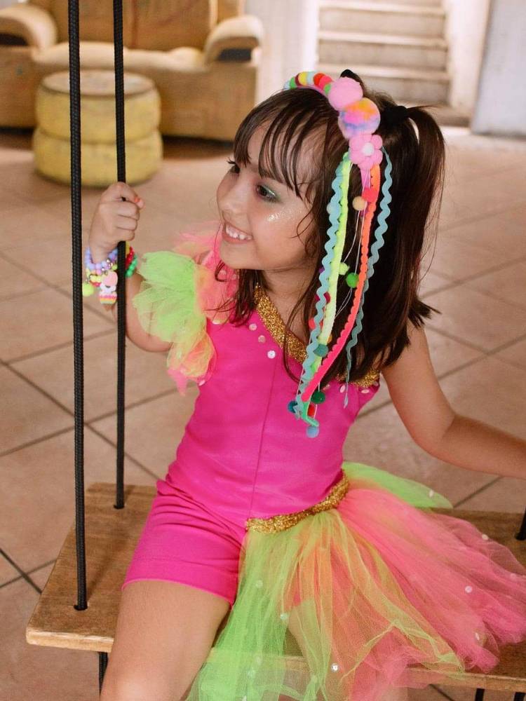 Criança de pele clara e cabelo castanho usando fantasia rosa com saia colorida e tiara de pompom em rosa, vermelho, azul e amarelo.