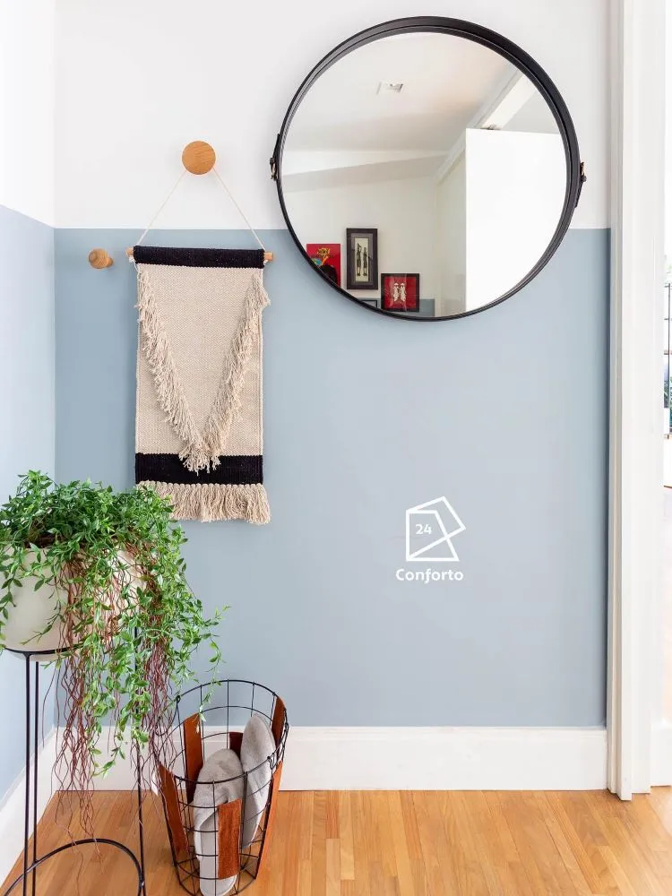 Parte de uma parede pintada com Conforto e a outra parte em branco, com tapete macramê pendurado, vaso com planta suspensa, espelho redondo e cesto vazado com chinelos dentro. 