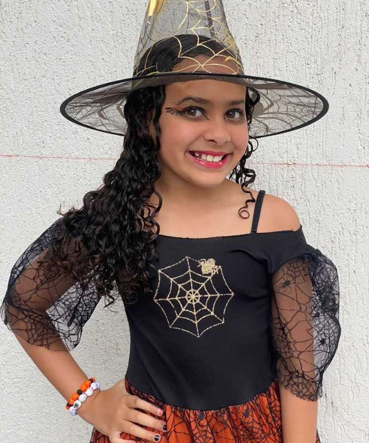 Garota fantasiada para o Halloween: Cores predominantes:preto e dourado . Usa: chapéu transparente pontudo, vestido, maquiagem de teia de aranha