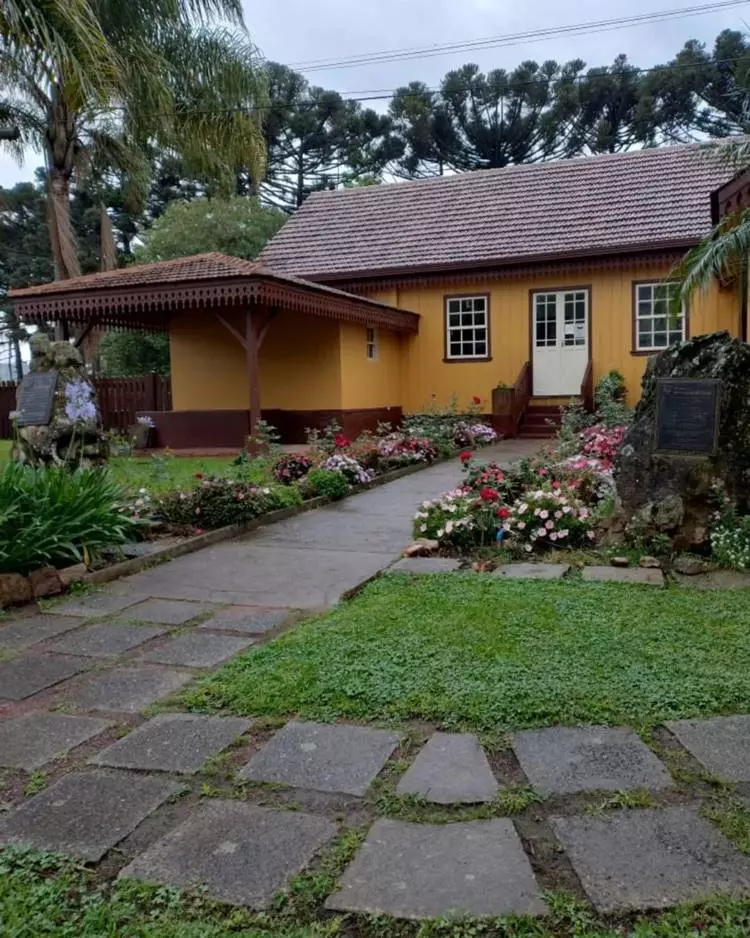 Imagem de fachada de casa simples amarela. Caminho de pedras rodeado de flores coloridas, rosa, branca, vermelha