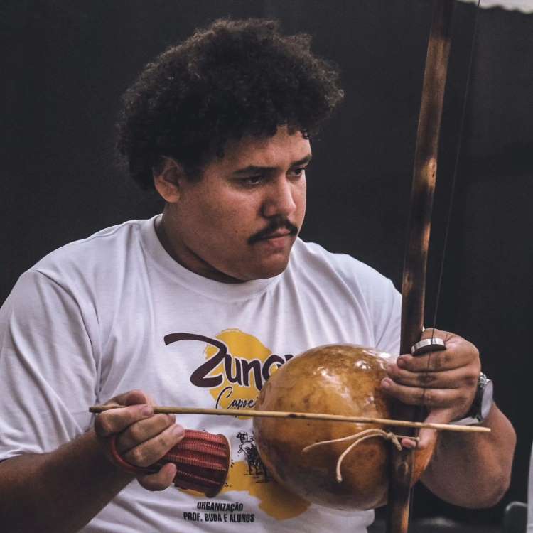 Lucas Henrique de blusa branca tocando berimbau. 