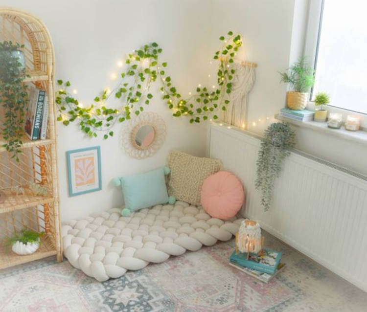 Cantinho com almofadas para meditar, com colchão bege no chão, almofadas, fio com luzes e folhas artificiais, estante de madeira, plantas, livros e janela com bastante iluminação.