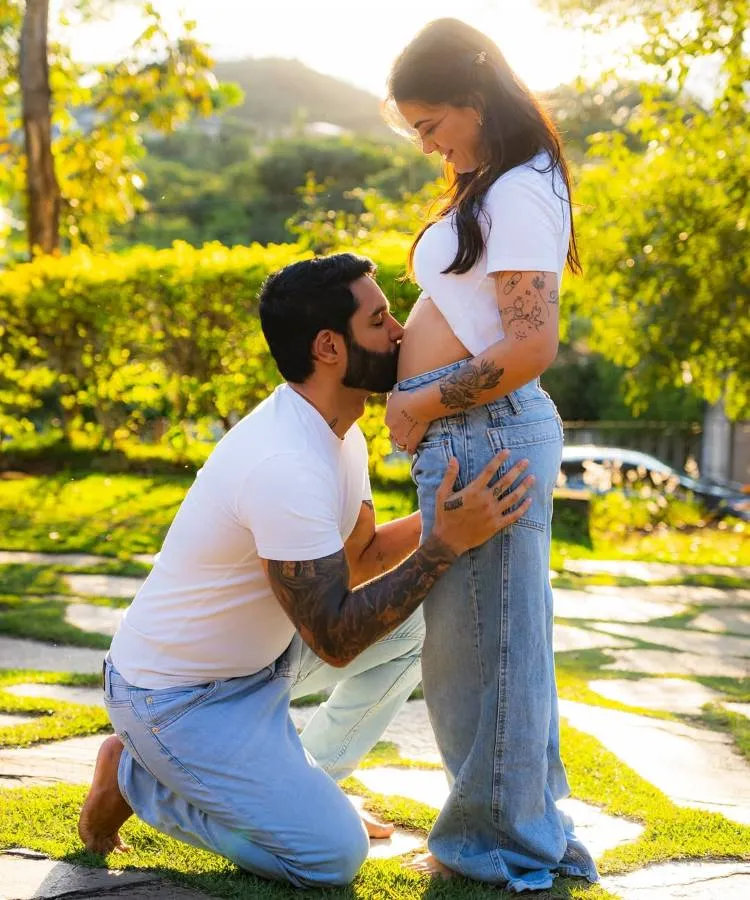 Casais famosos que reataram: Lucas Lira beija a barriga de Sunaika Bruna em uma foto ao ar livre. Ambos usam camisetas brancas e jeans claros. A imagem transmite carinho e a expectativa da gravidez.

