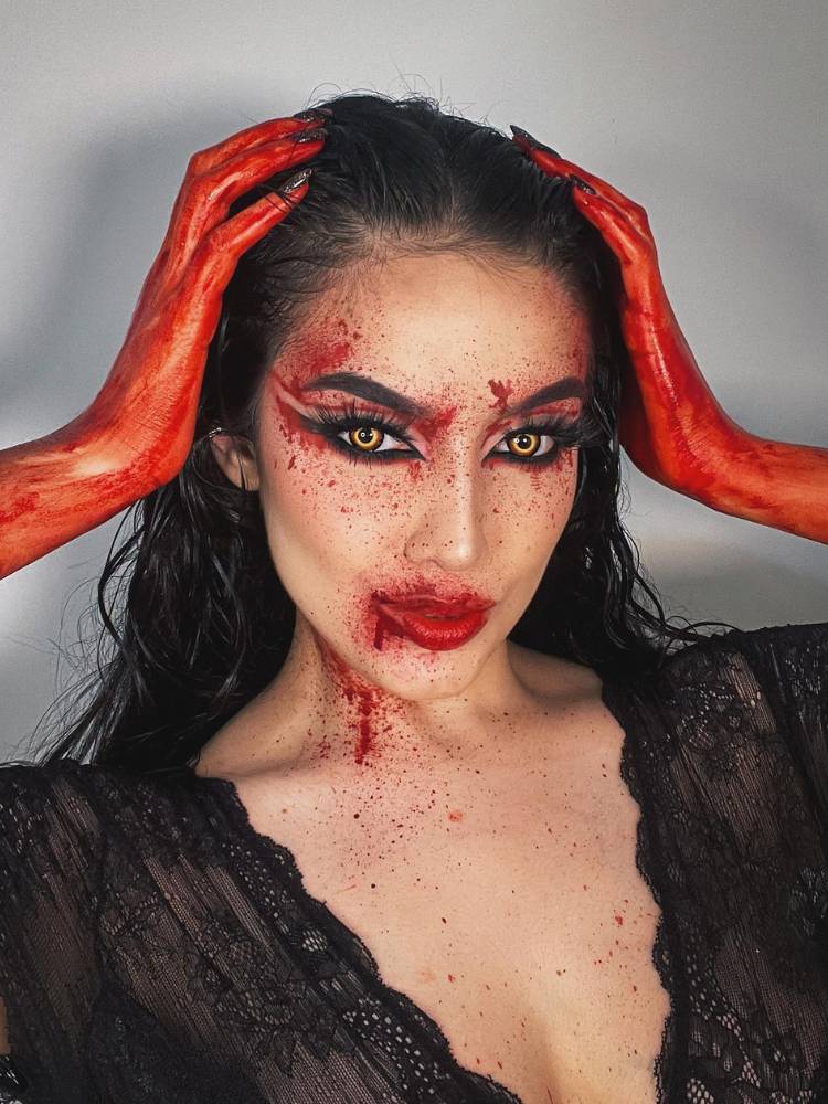 Mulher de pele clara com maquiagem vermelha com delineado, sangue artificial na boca, pescoço e mãos