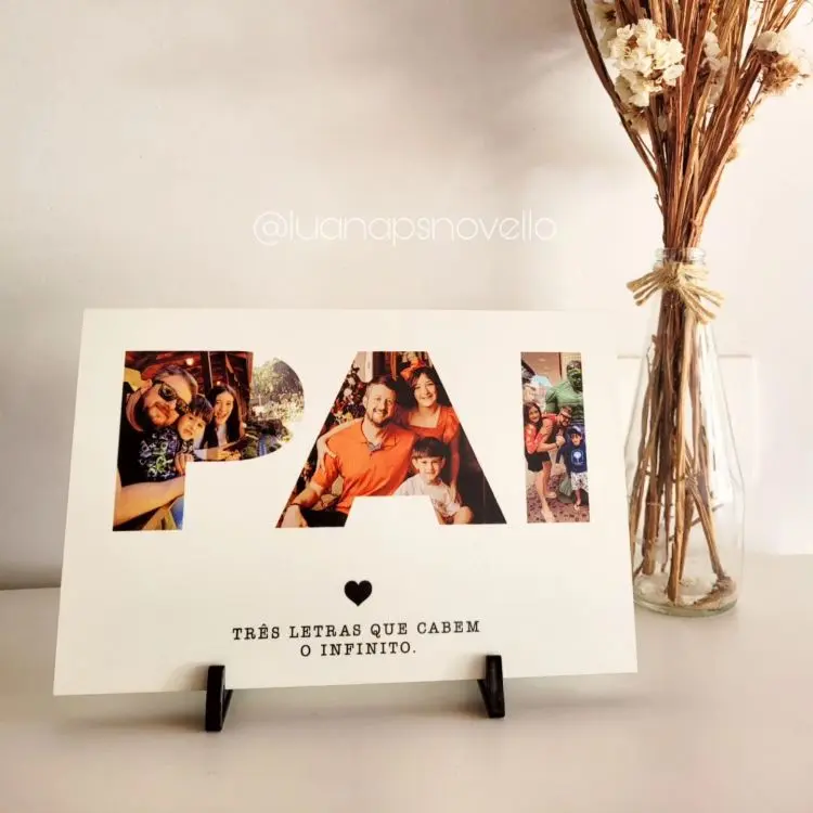 Porta-retato com recorte de letras formando a palavra “PAI”, preenchidas com fotos. Texto abaixo diz “TRÊS LETRAS QUE CABEM O INFINITO.” Apoiado em suporte, ao lado de flores secas.