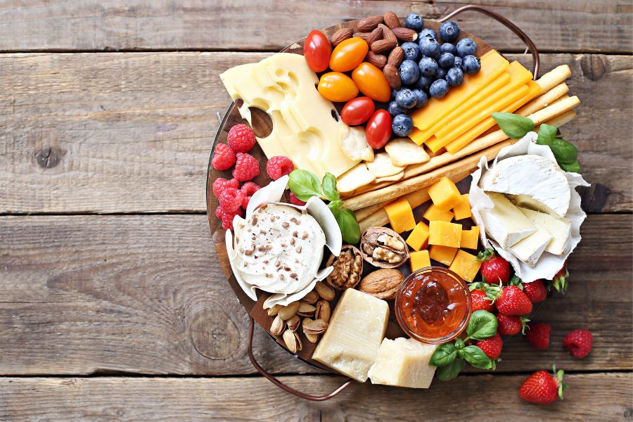 Tábua redonda de madeira com queijos brie, cheddar, gouda e parmesão, torradas, frutas, pistaches, nozes, amêndoas e geleia