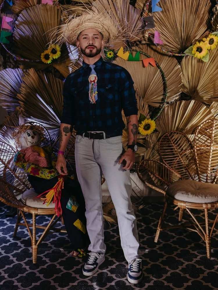 Homem de pele clara de camisa xadrez azul, calça e acessórios juninos