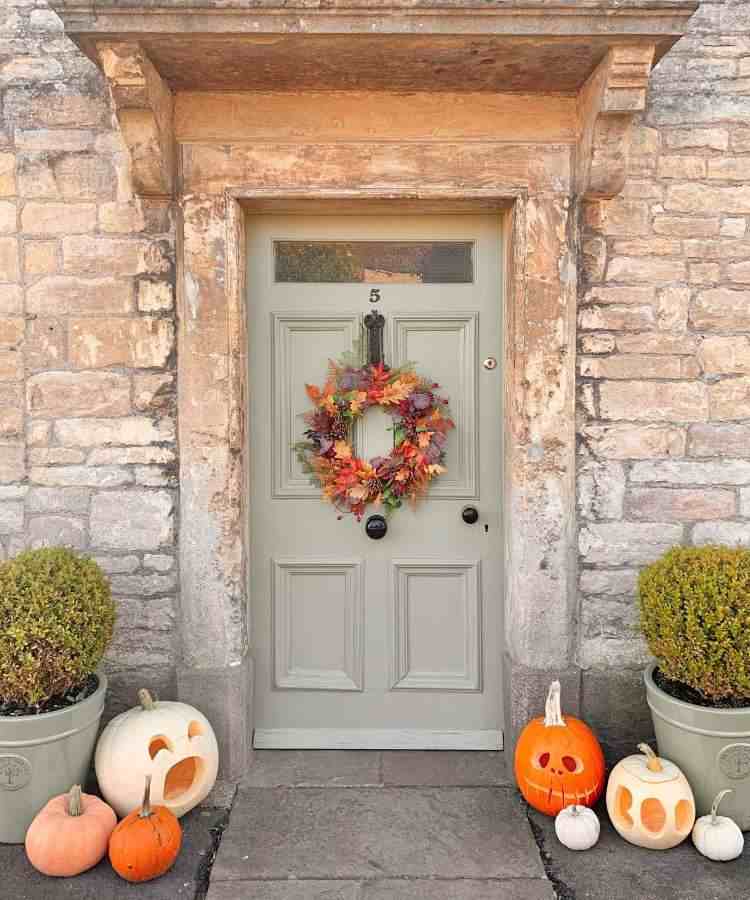 Decoração de entrada com tijolos à vista, porta cinza com guirlanda de flores alaranjadas, dois vasos com planta e abóboras recortadas 