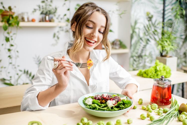 A imagem mostra uma pessoa sentada à mesa, desfrutando de uma refeição saudável. Ela está usando uma camisa branca e segurando um garfo com um pedaço de comida nele. A mesa é clara e há vários vegetais espalhados sobre ela, incluindo brócolis e cebolinhas. Há um pote de vidro contendo um líquido vermelho, possivelmente chá ou suco, ao lado do prato. O ambiente é iluminado e arejado, com plantas ao fundo e itens de cozinha visíveis nas prateleiras. A imagem transmite uma sensação de estilo de vida saudável e consciente.