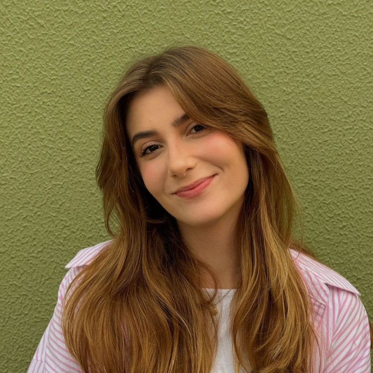 Jhenny Keller  com cabelos castanhos longos e soltos sorri suavemente. Ela veste camiseta branca e camisa listrada rosa. O fundo é uma parede verde texturizada, criando um contraste harmonioso com seu visual descontraído e alegre.