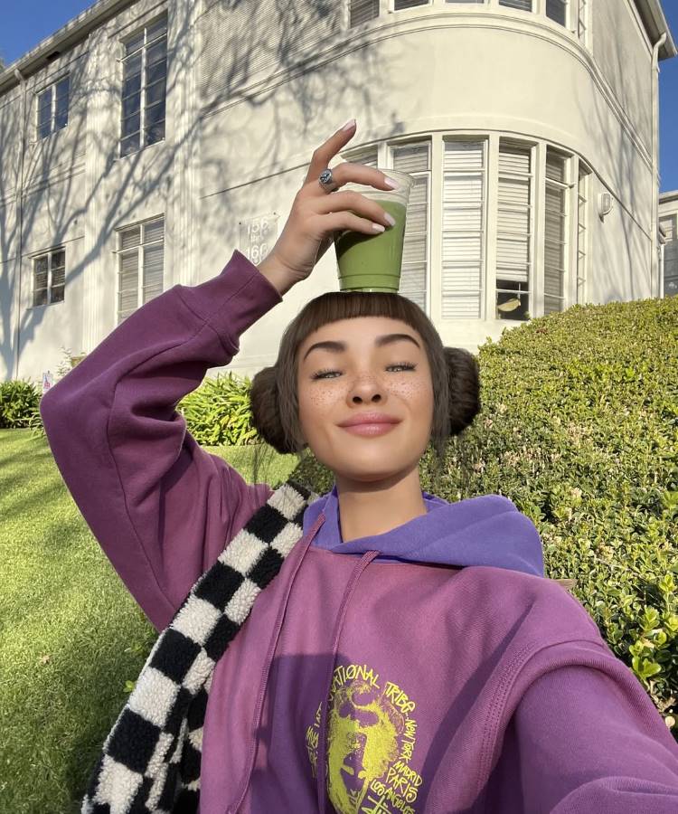 Uma jovem com penteado único em coques laterais e franja reta, possivelmente um dos influenciadores de IA, equilibra um copo de bebida verde na cabeça. Ela veste um casaco roxo e branco xadrez. Ao fundo, uma casa branca de múltiplos andares e vegetação sob um céu azul claro.