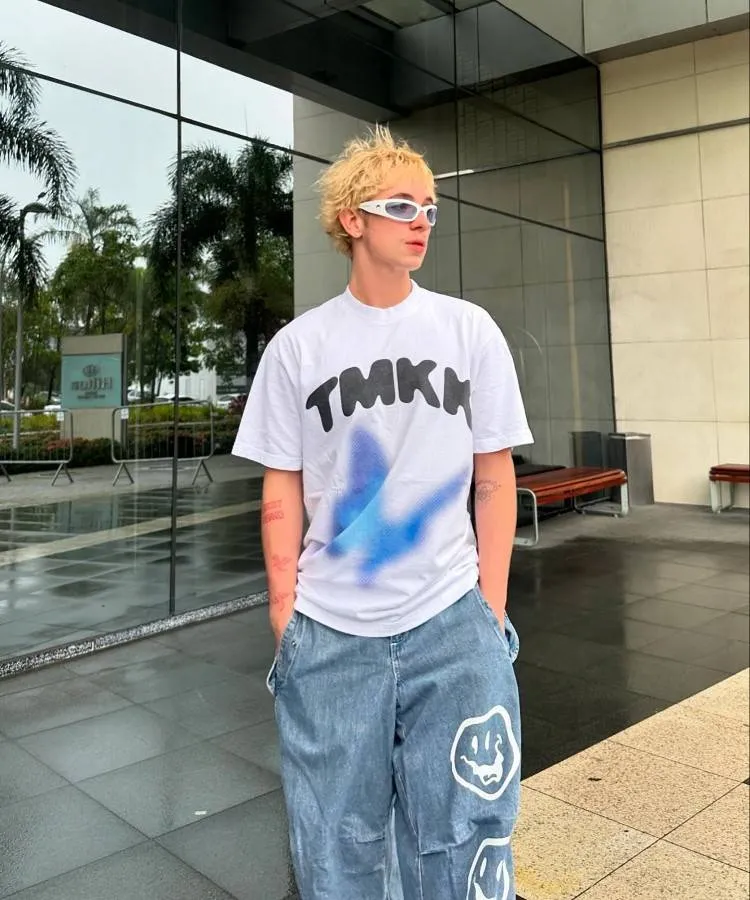 Juliano Floss usando camiseta, calça jeans e óculos em frente a um lugar com paredes de vidro