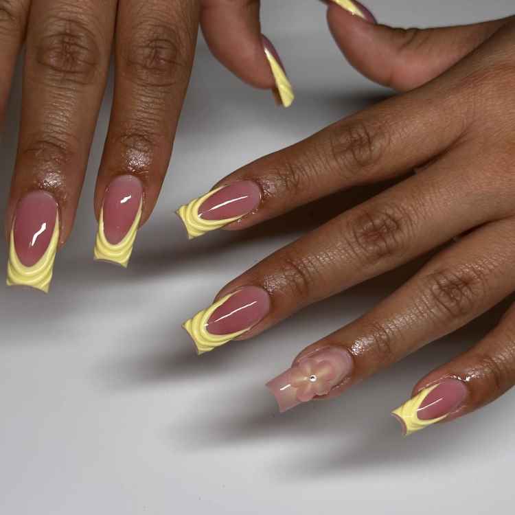 Unhas longas com esmalte rosa e decoração nas pontas em amarelo ondulado, criando um efeito tridimensional. Uma unha exibe um laço delicado, acentuando o visual criativo. As mãos repousam sobre um fundo branco, destacando o design único e moderno das unhas.