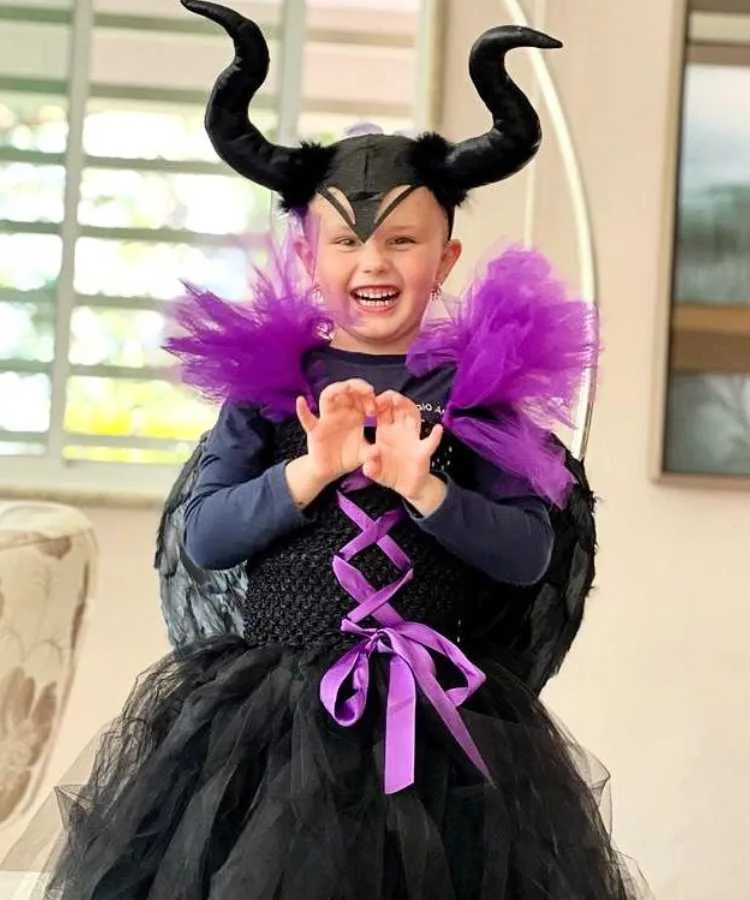 Garota com fantasia de bruxa infantil Malévola. Cores predominantes: . Usa: vestido + tiara de chifres da personagem + asas