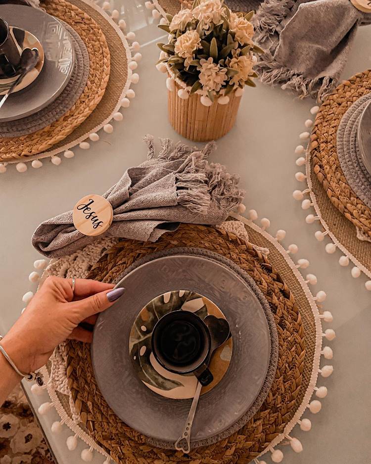 Uma mesa de café da manhã decorada para a Páscoa Cristã: sousplat boho, conjunto de xícaras cinza com estampa de costela de Adão, talheres em prata, centro de mesa com flor e guardanapo de tecido com prendedor de madeira escrito "Jesus".