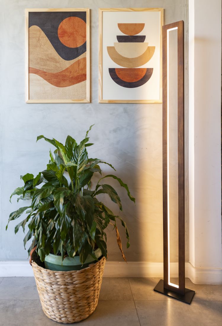 A imagem mostra um canto de sala decorado com uma planta em vaso de cerâmica verde, envolto por um cesto de palha, ao lado de uma luminária de piso moderna com estrutura de madeira e luz suave. Na parede de fundo, dois quadros abstratos com formas geométricas e tons terrosos contrastam com a parede de cimento queimado, criando um ambiente contemporâneo e acolhedor. A presença da planta traz frescor e vida ao espaço, exemplificando como decorar apartamento com plantas pode adicionar um toque natural e equilibrado à decoração. A combinação de elementos naturais e modernos cria uma atmosfera harmoniosa e convidativa.


