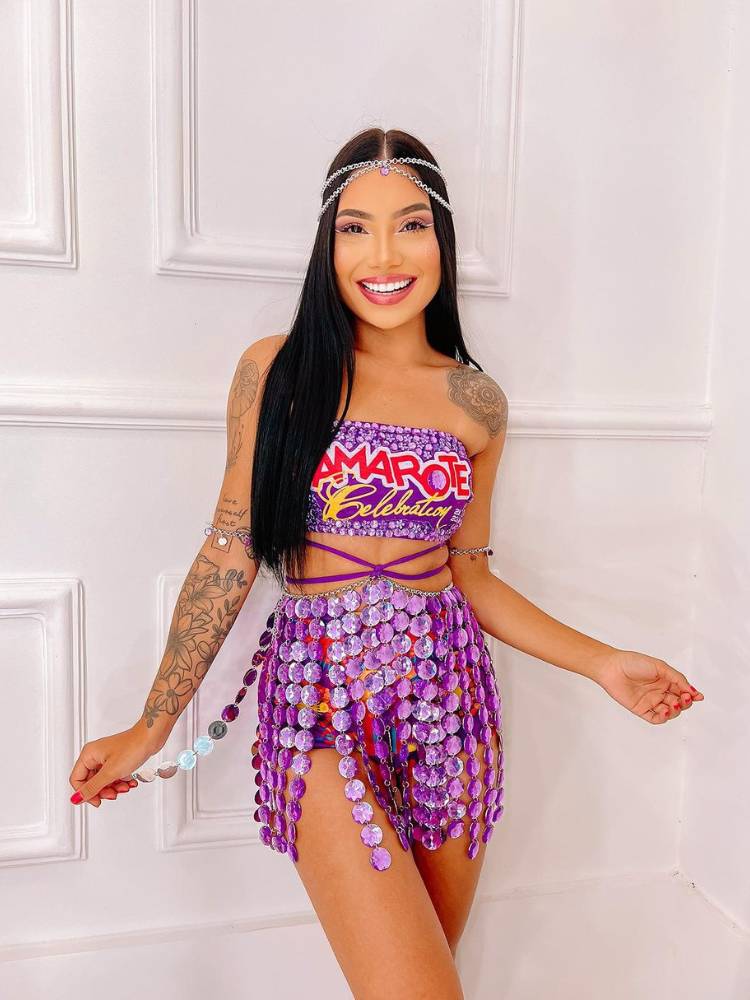 Mulher de pele clara e cabelo escuro liso usando brilho na cabeça, abadá customizado roxo em top com tiras e saia de brilho roxo. 