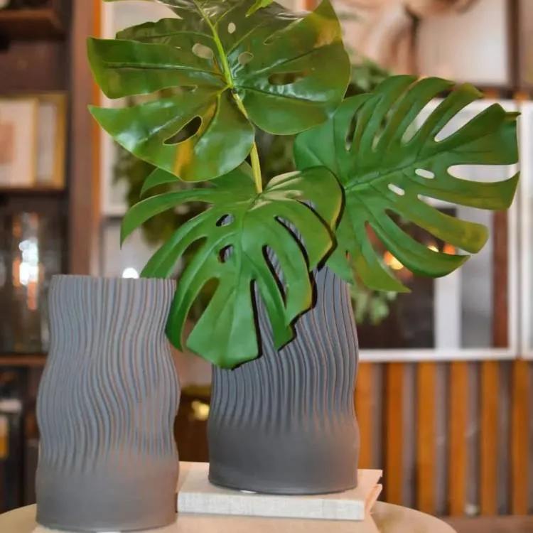 À esquerda, vaso cinza alto vazio. À direita, vaso cinza com folhas de costela-de-adão. 