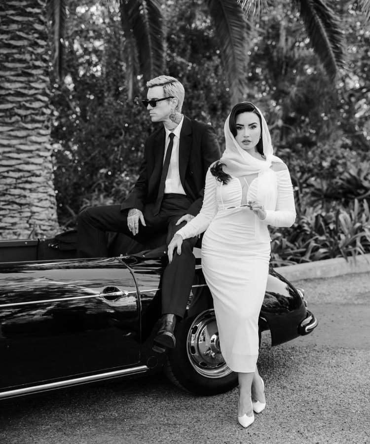 Em uma imagem em preto e branco, Demi Lovato, com lenço na cabeça e óculos de sol, posa elegantemente ao lado de um carro. Um homem de terno e óculos de sol está sentado na traseira do veículo, olhando para o lado, ambos em um cenário exterior com palmeiras.