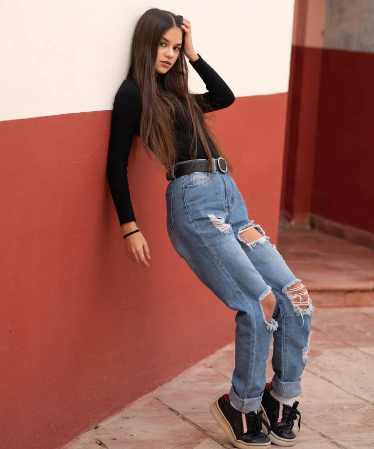 A imagem apresenta uma mulher vestindo uma calça jeans de cintura alta com rasgos marcantes, tendência 2025, combinada com um cinto preto e blusa de manga longa ajustada. O look é finalizado com tênis descolados, criando um estilo casual moderno.