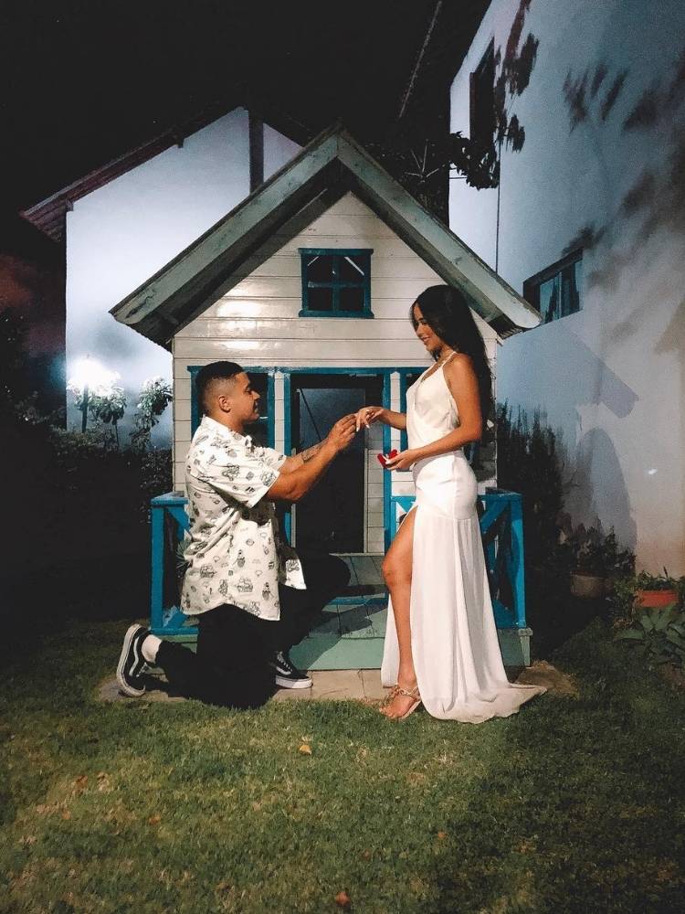 Michael Gomes usando camisa branca e calça preta ajoelhado pedindo a mão de Laura em casamento, que usa vestido longo branco. Ao fundo, xalé branco
