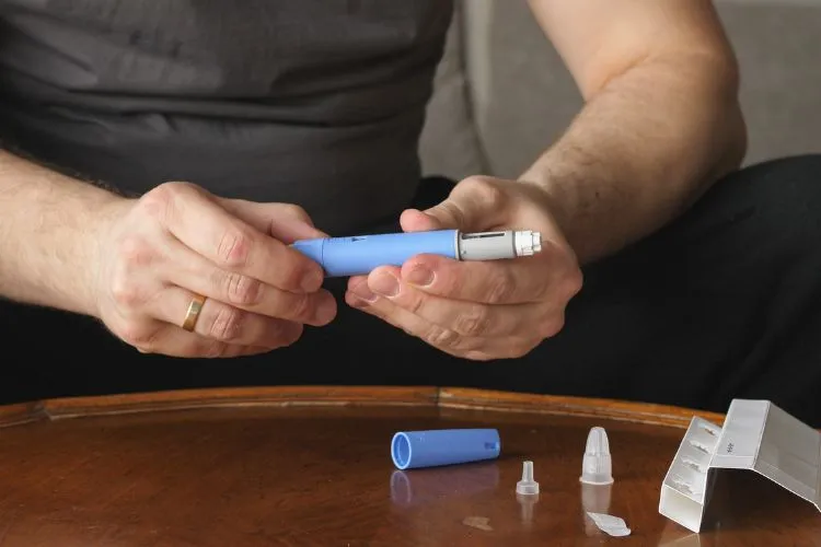 Homem segurando uma caneta aplicadora azul clara de Mounjaro, preparando-se para injeção, sentado diante de uma mesa de madeira com acessórios médicos próximos. Foca-se nas mãos, mostrando cuidados durante a manipulação do medicamento.