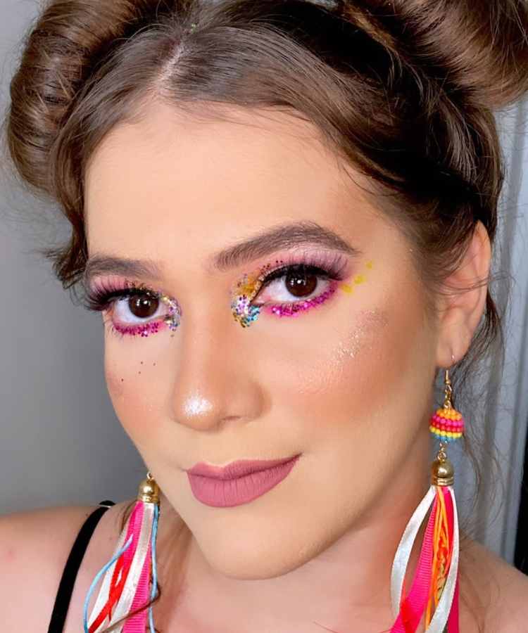 Mulher de pele clara com space buns e olho rosa com glitter colorido, além de brincos coloridos
