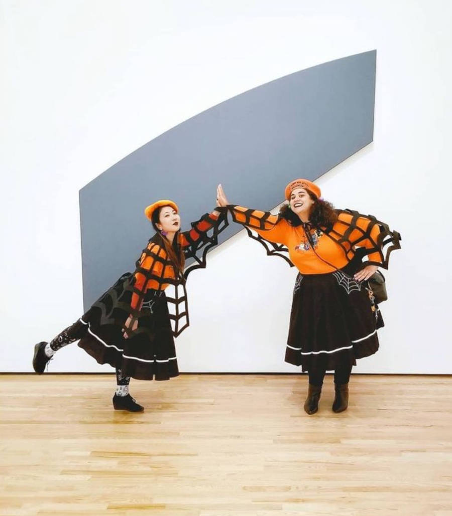 Imagem de duas mulheres distantes encostando as mãos vestidas com fantasia de Halloween composta com a capa de Teia de Aranha, saia preta e blusa e boina laranjas 