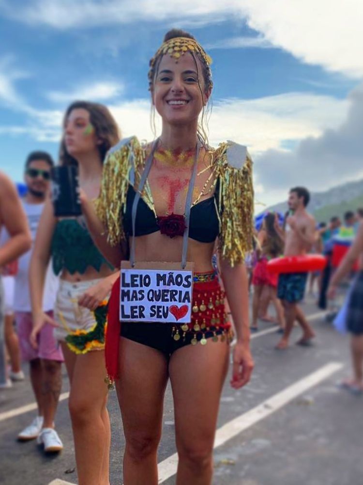 Mulher de pele clara no carnaval usando top preto, ombreira de fitas coloridas, hot pant preta, lenço vermelho, acessórios que imitam moedas e plaquinha "leio mentes mas queria ler seu (desenho de coração".
