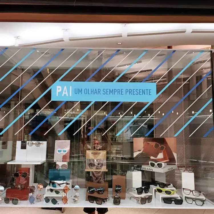 A imagem exibe a vitrine de uma ótica, decorada com a frase "PAI UM OLHAR SEMPRE PRESENTE" em destaque, sobreposta a linhas diagonais azuis e brancas. A vitrine está repleta de óculos de sol e de grau em expositores, com algumas imagens de modelos usando-os.
