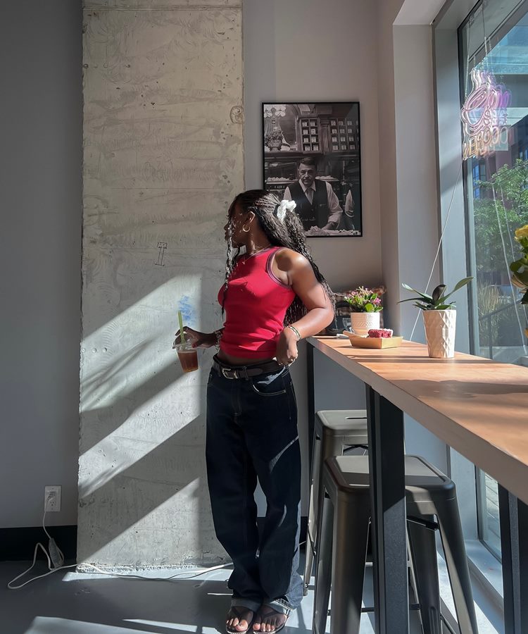 Uma jovem mulher com longas tranças e uma flor branca no cabelo, usando uma regata vermelha e jeans largos, está de pé em um café. Ela segura um copo e observa pela janela ensolarada. A cena captura um momento relaxante e casual.