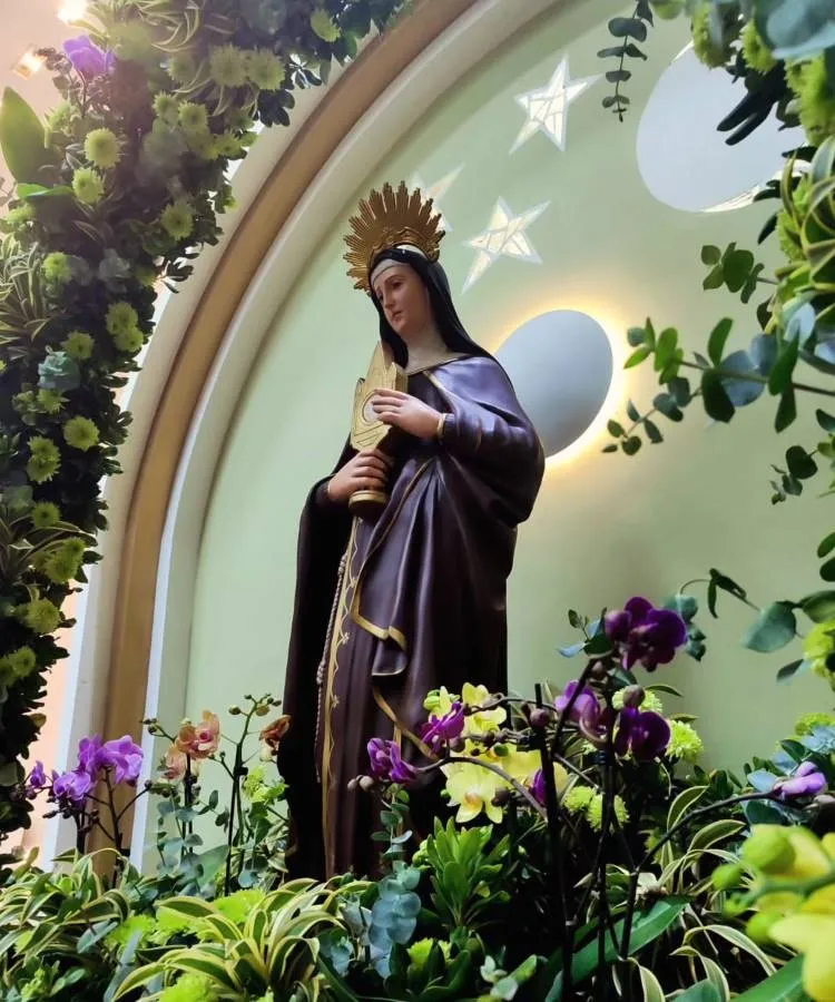 A imagem mostra Santa Clara de Assis em um hábito marrom com detalhes dourados e véu preto. Ela segura um ostensório dourado e usa uma auréola dourada com raios. A estátua está cercada por flores coloridas, com um fundo verde claro e estrelas brancas, criando um ambiente celestial.




