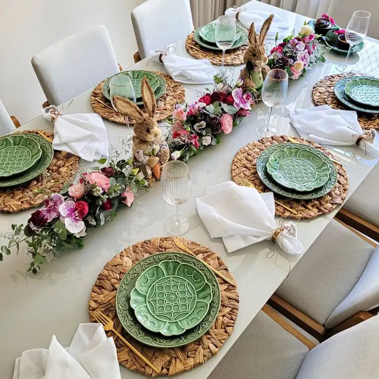  mesa posta de Páscoa com centro floral e coelhos, sousplats de palha. pratos verdes com taças de vidro
