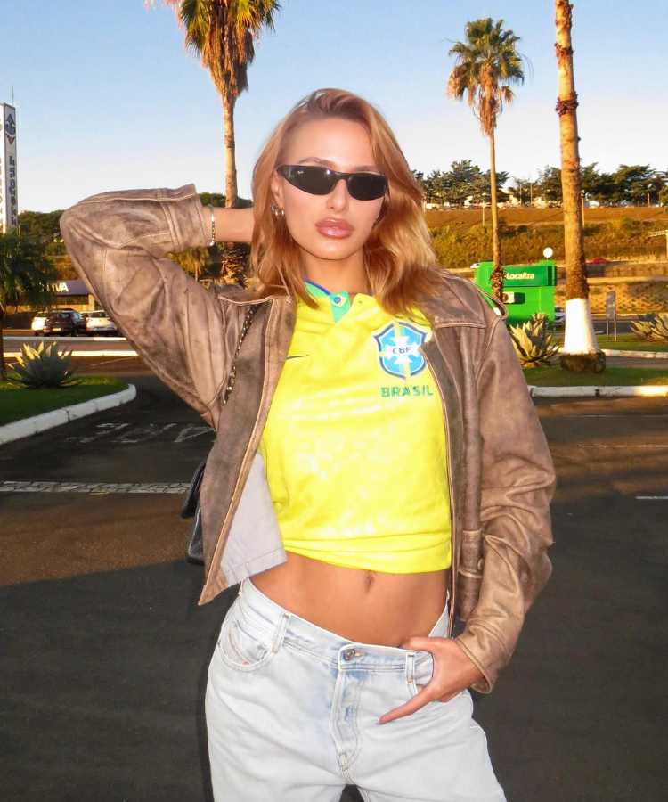 Imagem com fundo de rua com carros, palmeiras. No centro , mulher usando camisa do Brasil + jaqueta de couro + calça jeans