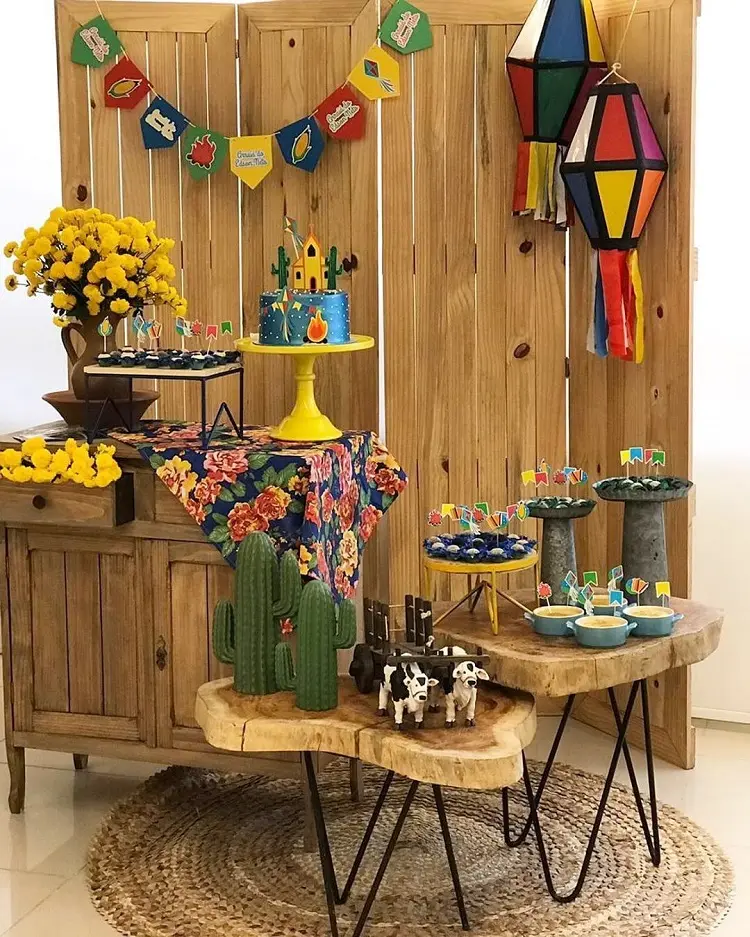 A imagem representa decoração para festa junina com mesas diferentes, em formato móvel de madeira. Contém doces, cactos, flores, balões, bandeirinhas, etc