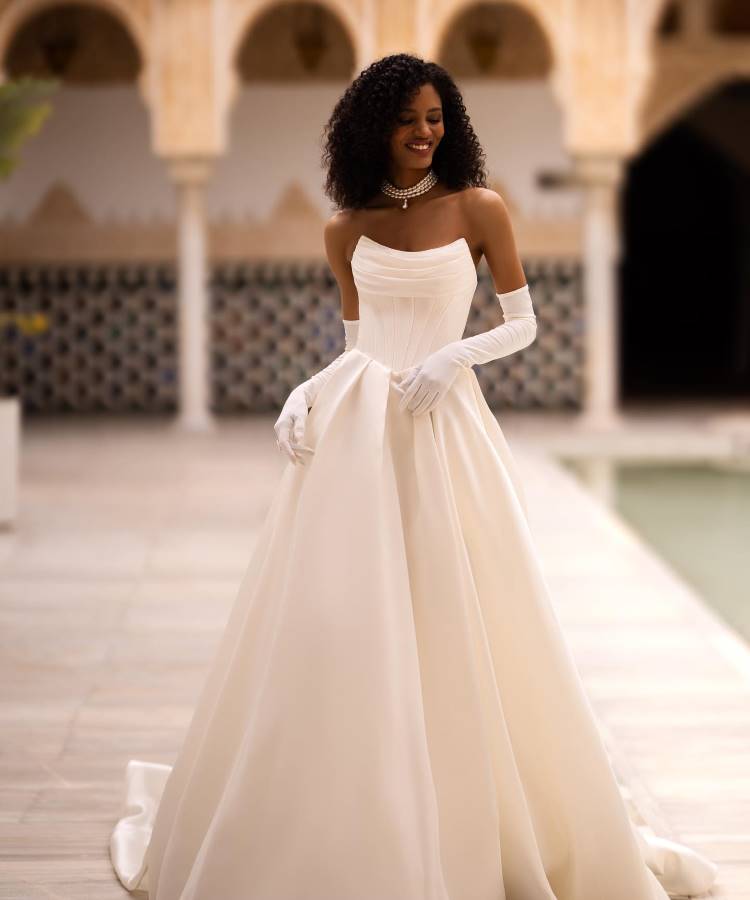 Noiva em vestido branco clássico com corpete estruturado, decote reto e saia volumosa, complementado por luvas longas e colar de pérolas. O design elegante reflete as tendências em vestidos de noiva 2025, em cenário com arquitetura árabe sofisticada.