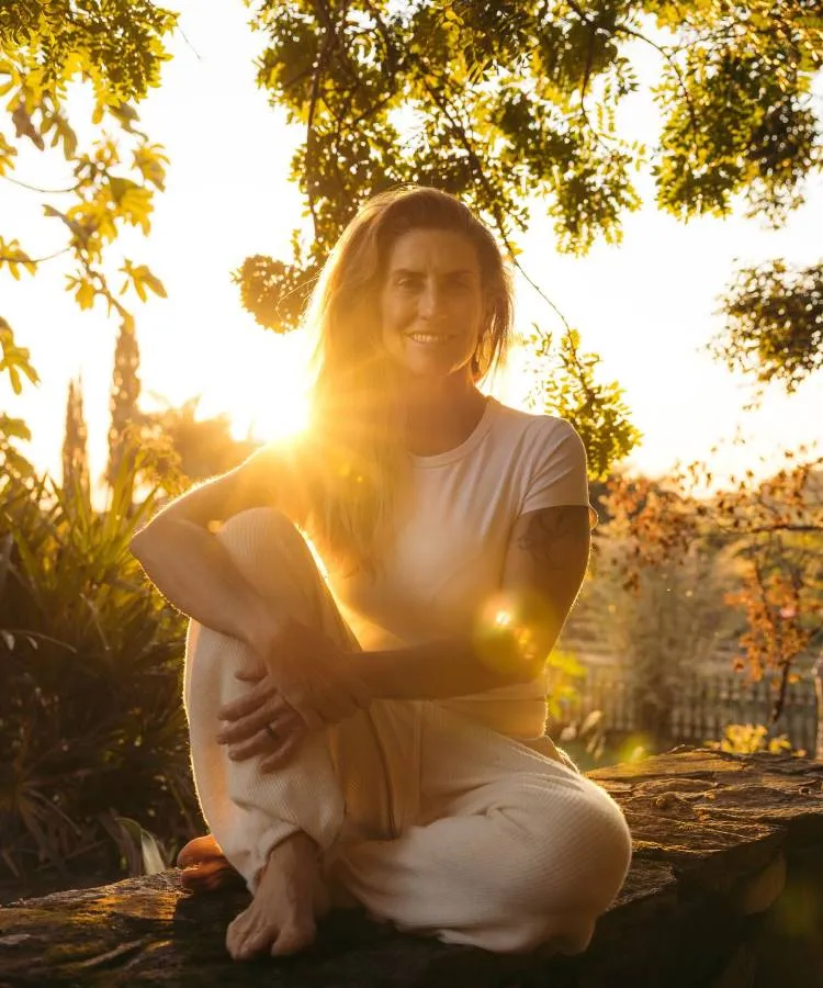 A imagem mostra Alessandra Luglio sorridente sentada em um ambiente externo, iluminada por um sol intenso vindo de trás. Ela veste roupas claras e parece relaxada, com árvores e folhagens desfocadas ao fundo, criando uma atmosfera serena e natural.