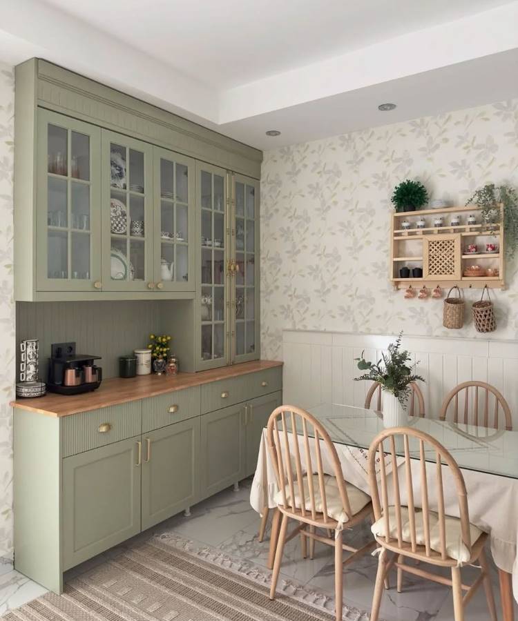 Cozinha com decoração Feng Shui, destaca móveis verdes em madeira clara e bancada organizada. Mesa de vidro adornada com toalha neutra, cadeiras de madeira e flores naturais. Estante delicada e papel de parede floral criam harmonia e energia fluida.





