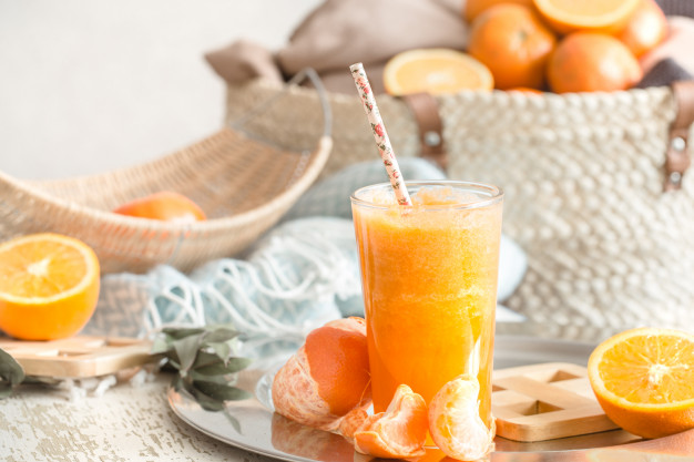 Foto de suco de laranja com laranjas e mexericas ao redor, além de cestos de palhas com essas frutas ricas em vitamina C 