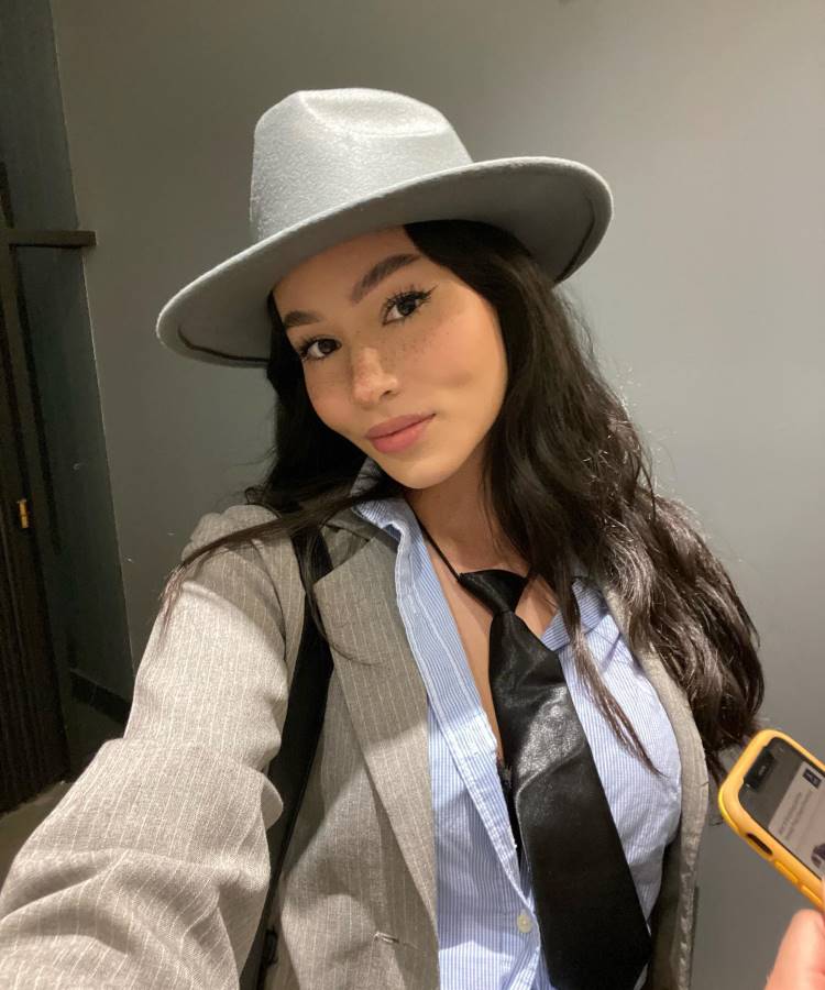 A imagem mostra uma pessoa vestida com uma fantasia inspirada em Oppenheimer. Ela está usando um chapéu cinza e um terno listrado, com uma camisa azul clara e uma gravata preta solta. O cabelo é longo e solto, e a expressão é confiante e sorridente. Ela segura um celular com capa amarela na mão direita. O fundo é neutro, destacando a roupa e o estilo da fantasia.


