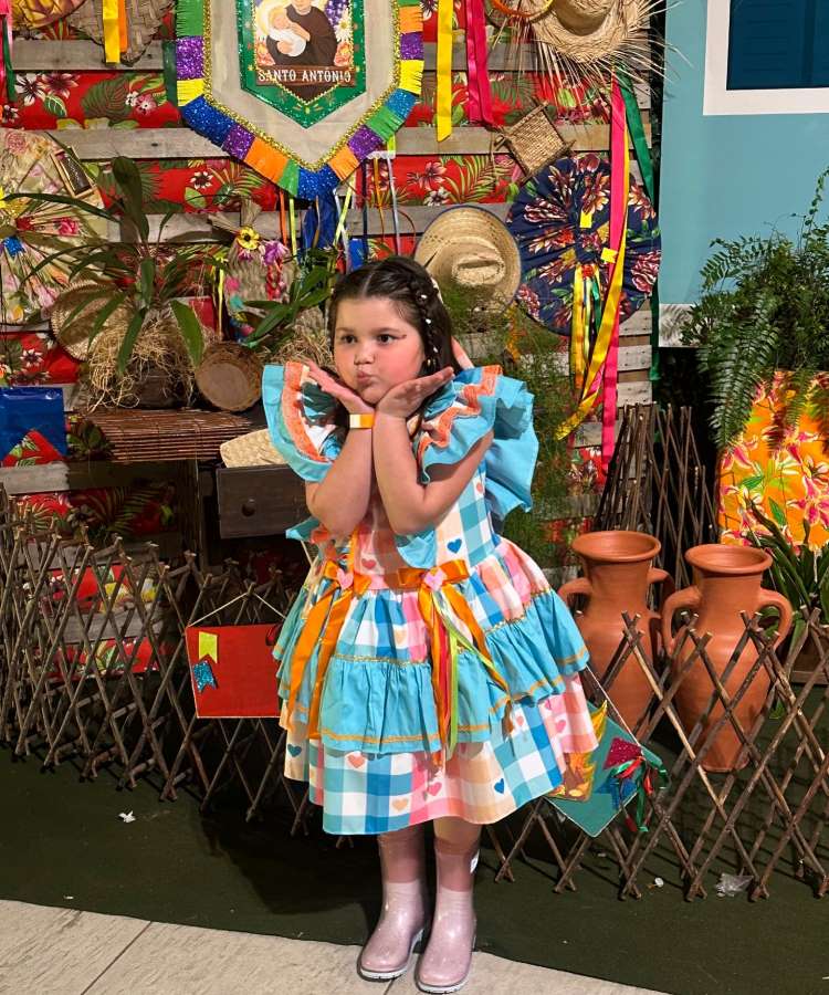 Menina usando vestido de quadrilha colorido, botas transparentes, com fundo de painel decorado.
