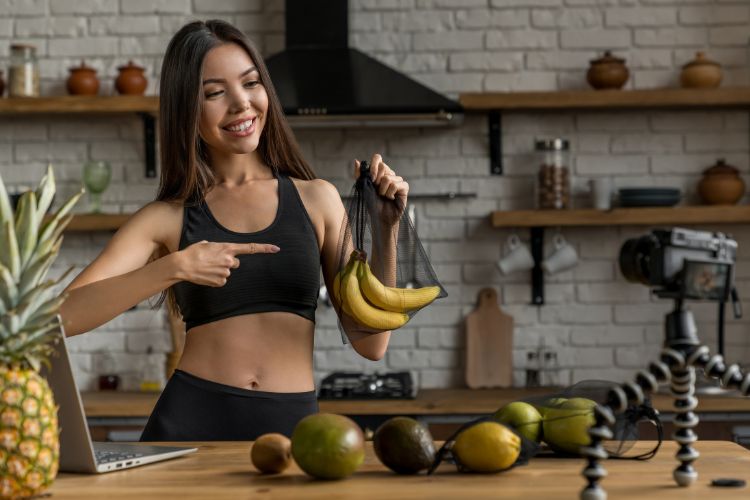 Mulher sorridente em cozinha moderna aponta para bananas em uma sacola reutilizável. Ela veste roupa fitness preta e filma conteúdo com câmera sobre tripé. Frutas tropicais, como abacaxi e limão, estão sobre a mesa ao lado de laptop.